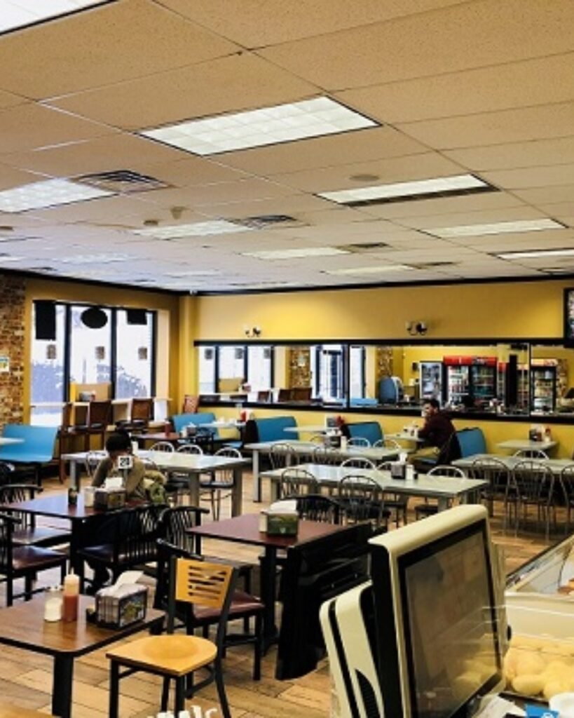 Cafeteria com mesas e cadeiras, balcão de atendimento ao fundo e algumas pessoas sentadas