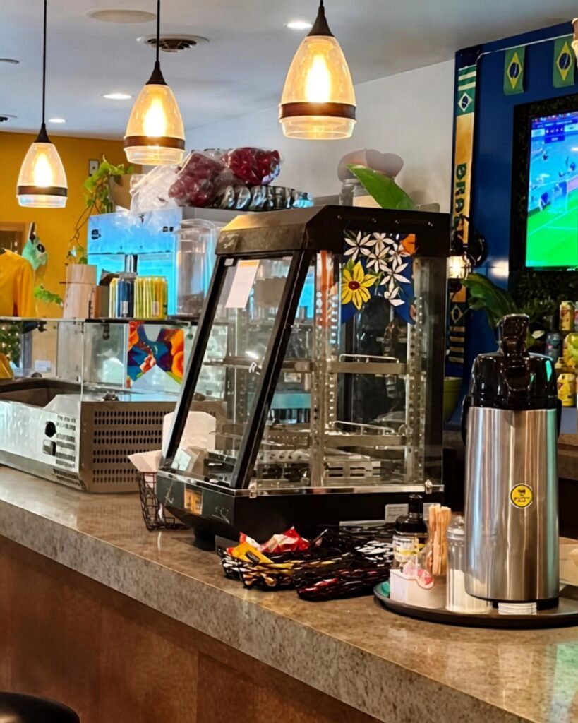 Balcão de lanchonete com estufa de alimentos, bebidas, luminárias pendentes e televisão exibindo um jogo de futebol