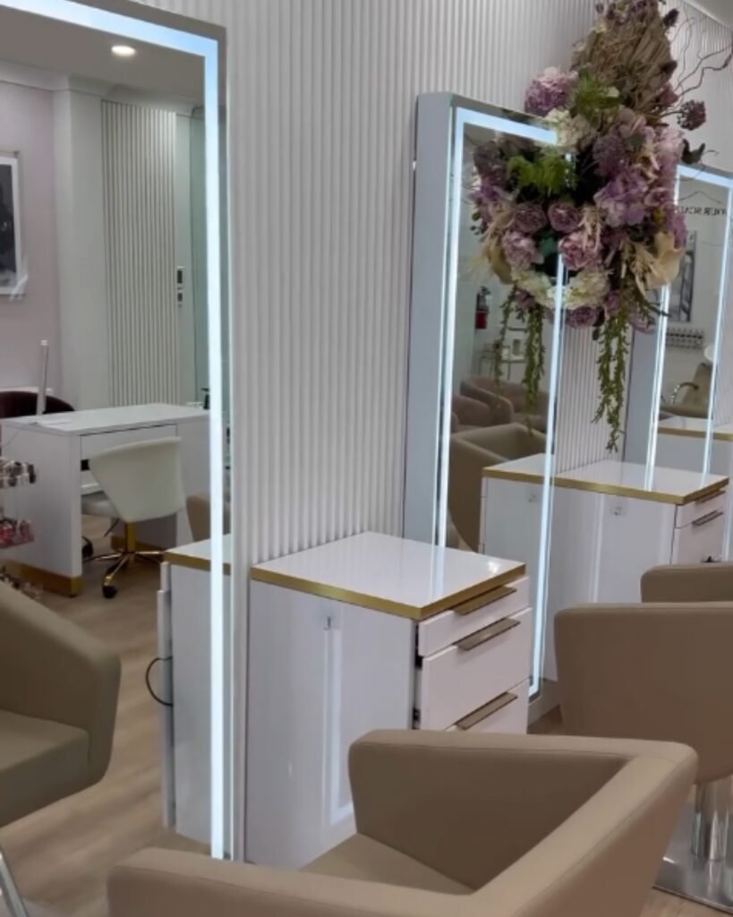 Interior de um salão de beleza com espelhos iluminados, bancadas brancas, poltronas e arranjo floral